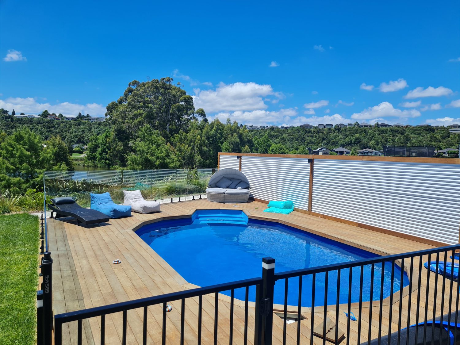 A pool in a timber deck behind a black fence, with water and trees beyond