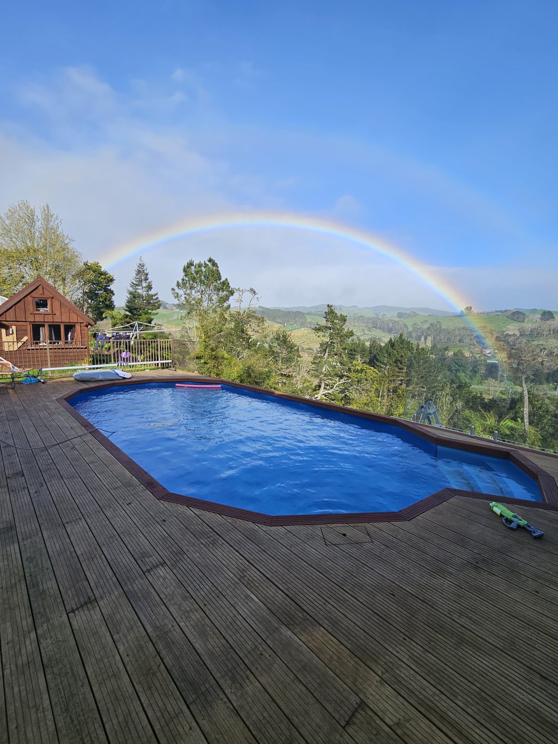 A pool set into a dark timber deck with a rainbow over the hills behind